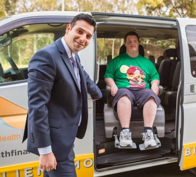 William Johns smiling beside HFI’s wheelchair-accessible van, with a client seated in his wheelchair inside the vehicle